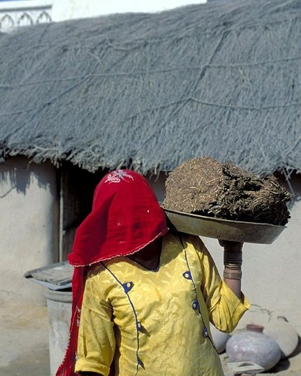 Woman in a red veil and yellow dress carrying a load on her head outside a thatched house